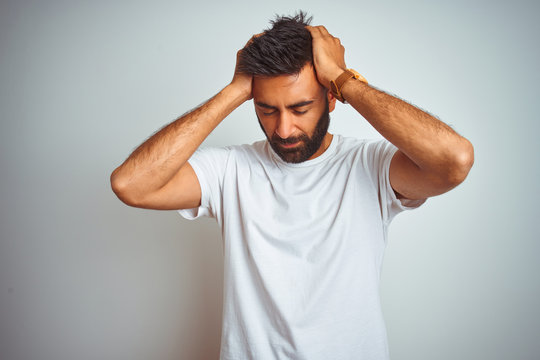 Young indian man wearing t-shirt standing over isolated white background suffering from headache desperate and stressed because pain and migraine. Hands on head.