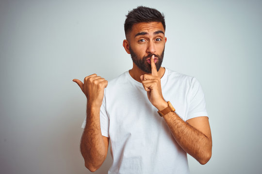 Young indian man wearing t-shirt standing over isolated white background asking to be quiet with finger on lips pointing with hand to the side. Silence and secret concept.
