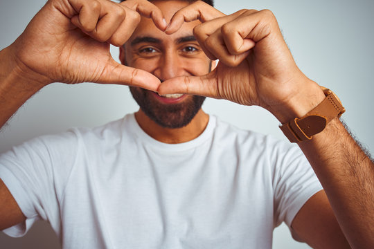 Young indian man wearing t-shirt standing over isolated white background smiling in love showing heart symbol and shape with hands. Romantic concept.