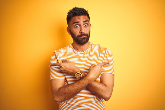 Young indian man wearing t-shirt standing over isolated yellow background Pointing to both sides with fingers, different direction disagree
