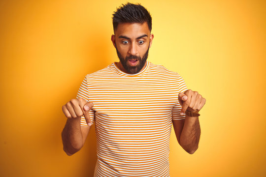 Young indian man wearing t-shirt standing over isolated yellow background Pointing down with fingers showing advertisement, surprised face and open mouth