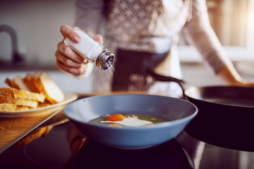 Close up of caucasian woman adding salt in sunny side up eggs while standing in kitchen next to stove. - Powered by Adobe