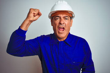 Handsome middle age worker man wearing uniform and helmet over isolated white background angry and mad raising fist frustrated and furious while shouting with anger. Rage and aggressive concept.