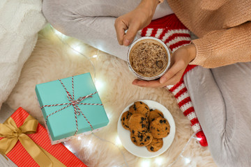 Woman with cup of hot drink, cookies and gifts on white fuzzy carpet, top view. Christmas season