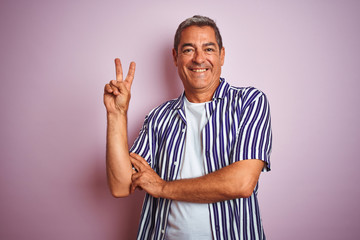 Handsome middle age man wearing striped shirt standing over isolated pink background smiling with happy face winking at the camera doing victory sign. Number two.