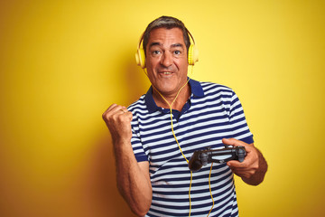 Middle age man playing video game using headphones over isolated yellow background screaming proud and celebrating victory and success very excited, cheering emotion