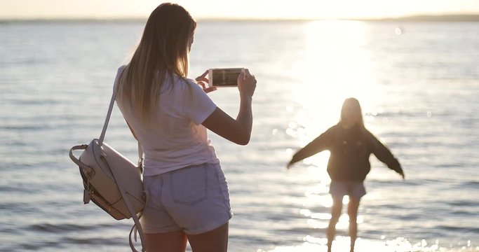 A young mother shoots a daughter on a mobile phone on the background of the sunset.