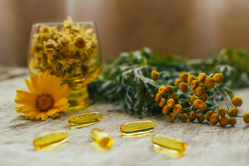 Obraz na płótnie Canvas Fish oil capsules, tansy, dry chamomile flowers in glass cup, calendula on linen fabric at the table, copy space