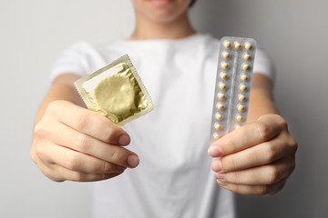 Young woman holding birth control pills and condom on light grey background, closeup. Safe sex...