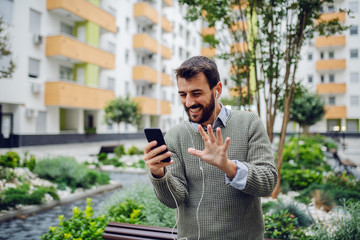 Laughing handsome caucasian fashionable businessman having video call over smart phone and waving while standing in park.