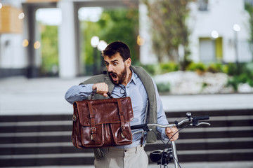 Good-looking fashionable caucasian businessman holding his briefcase, pushing bicycle, looking at...