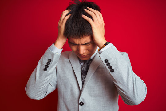 Asian chinese businessman wearing grey jacket and tie standing over isolated red background suffering from headache desperate and stressed because pain and migraine. Hands on head.