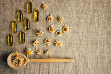 Layout with dry chamomile flowers in wooden spoon and fish oil capsule on linen fabric at the table, copy space