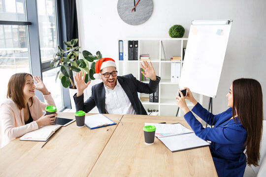 Workers in office. Celebrating new year or Christmas. People sit at table together. Brunette taking picture of man in red hat. Having fun at work.