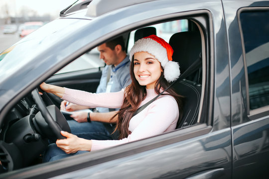 Young beautiful cheerful woman sit on driver's place in car. Holding hands on steering wheel and smile. Wear red hat. Christmas or new year period. Man sit besides her.
