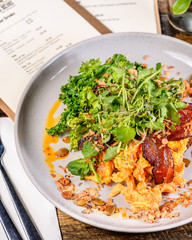 Scrambled Eggs with bacon, herbs and greens on a round plate with menus in the background.