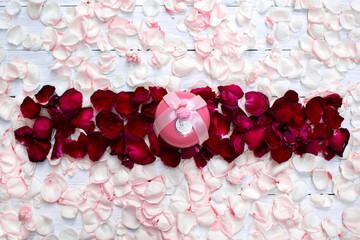 Pink circle gift box out standing from horizontal bar made of red rose petals with light pink rose petals scattered around on white wooden background. Top view. Valentine's background. Copy space.