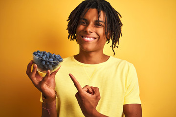 Afro american man with dreadlocks holding blueberries over isolated yellow background very happy pointing with hand and finger
