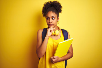 Afro woman using backpack and headphones holding notebook over isolated yellow background serious...