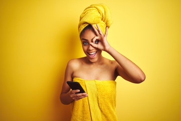 Afro woman wearing towel after shower using smatrphone over isolated yellow background with happy...