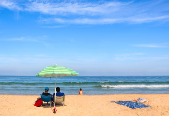 Woman and man sit on beach chairs opposite the sea under a striped sun umbrella . Young girl sits...