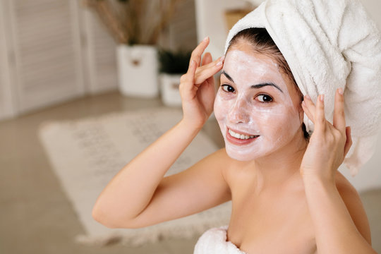smiling woman with towel is applying mask on her face and looking at the camera at home. Skin care. space for text