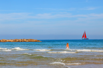Sunny calm sea seascape. Young woman in a swimsuit goes in the surf. Sailboat with a red sail on the horizon