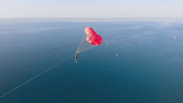 Towing a multicolored red parachute behind a boat jetski over the sea along the beach at sunset. Drone flied near the parachute. Aerial view.