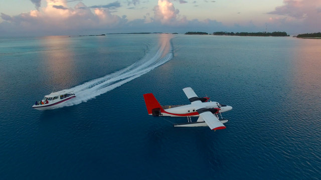 Aerial photo of seaplane during sunset. White black red seaplane docked in the middle of the Maldivian lagoon of Indian Ocean, far away from Island. Speed boat is passing by