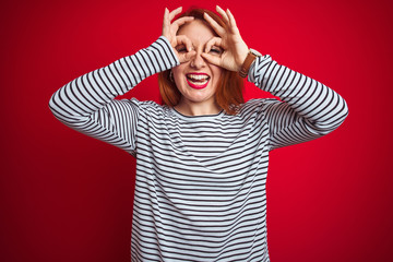 Young redhead woman wearing strapes navy shirt standing over red isolated background doing ok gesture like binoculars sticking tongue out, eyes looking through fingers. Crazy expression.