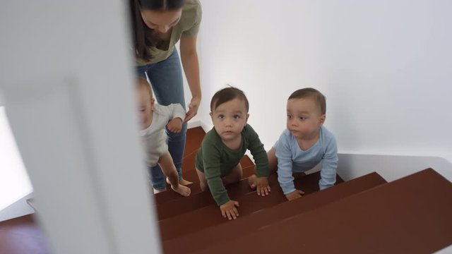Trucking shot of Asian toddler triplets in bodysuits climbing up staircase at home, and young mom helping boy walk up stairs