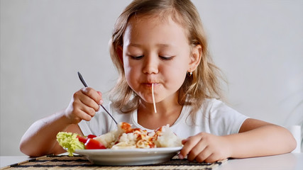 A little girl is eating spaghetti with vegetables at the table in the light room with a fork . Portrait of a kid eat at home in front of the camera.