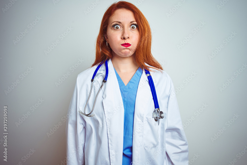 Sticker Young redhead doctor woman using stethoscope over white isolated background puffing cheeks with funny face. Mouth inflated with air, crazy expression.