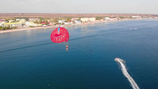 Towing a multicolored red parachute behind a boat jetski over the sea along the beach at sunset. Drone flied near the parachute. Aerial view.
