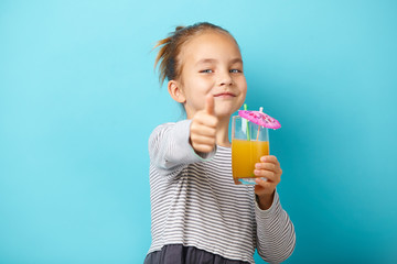 little child girl with orange juice, shows thumbs up, has a positive emotions, stands on blue backgound.