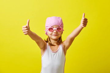 Little girl in a bathing suit shows thumbs up, wearing a swimsuit, swimming cap and glasses, stands...