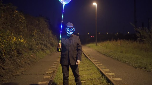Man with purge mask and balloon is standing on a railway at night.