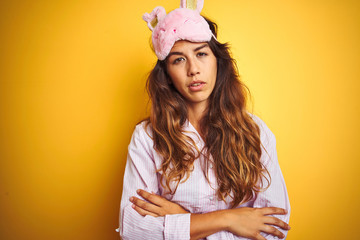Naklejka premium Young woman wearing pajama and sleep mask standing over yellow isolated background looking sleepy and tired, exhausted for fatigue and hangover, lazy eyes in the morning.