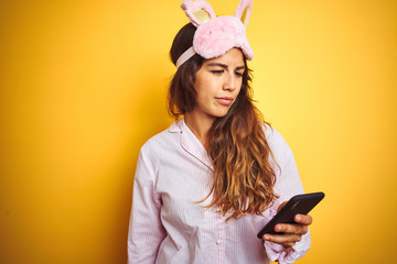 Fototapeta na wymiar Young woman wearing pajama and mask using smartphone over yellow isolated background with a confident expression on smart face thinking serious
