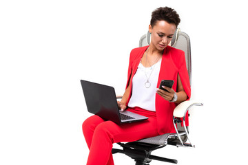 A young beautiful girl with short dark hair, makeup in a red office suit with bijuteria, expensive watches, sits in a computer chair with a laptop and a phone in hand