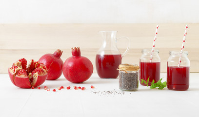 Pitcher and two jars of fresh pomegranate juice, with three ripe pomegranates, one freshly peeled. Rouge pomegranate seeds and chia seeds in the foreground, all on a white table.