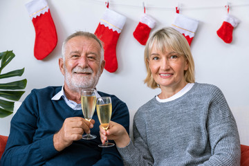 Senior couple man and woman with champagne and chatting, sitting on sofa with Christmas tree and decoration in background; looking at camera