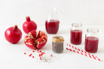 Pitcher and two jars of fresh pomegranate juice, with three ripe pomegranates, one freshly peeled. Rouge pomegranate seeds and chia seeds in the foreground, all on a white table.