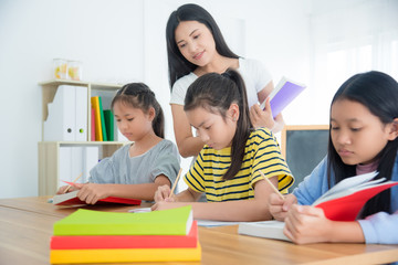 Three asian students sitting and reading book in the classroom with a teacher standing by.