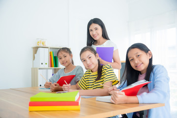Three asian students sitting and reading book in the classroom with a teacher standing by.