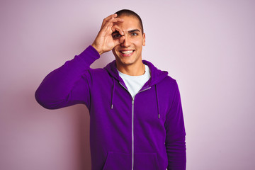 Young handsome man wearing purple sweatshirt over pink isolated background doing ok gesture with hand smiling, eye looking through fingers with happy face.