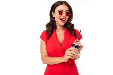 The young woman is red lips, bright makeup, dark wavy long hair, in a red suit, with red square glasses and a cup of coffee with a tube