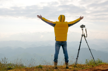 The photographer with yellow jacket stand near to his camera and stand in front cliff, cloudy and show action of happy.