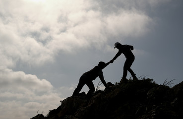 Silhouette image of one friend try to pull his other friend over the cliff of mountain and they climb and help each other during day time of travel.