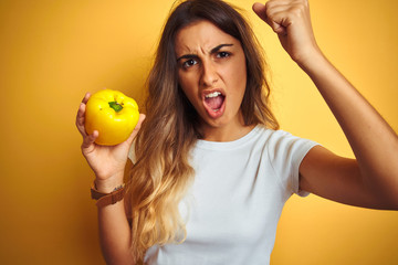 Young beautiful woman holding pepper over yellow isolated background annoyed and frustrated shouting with anger, crazy and yelling with raised hand, anger concept